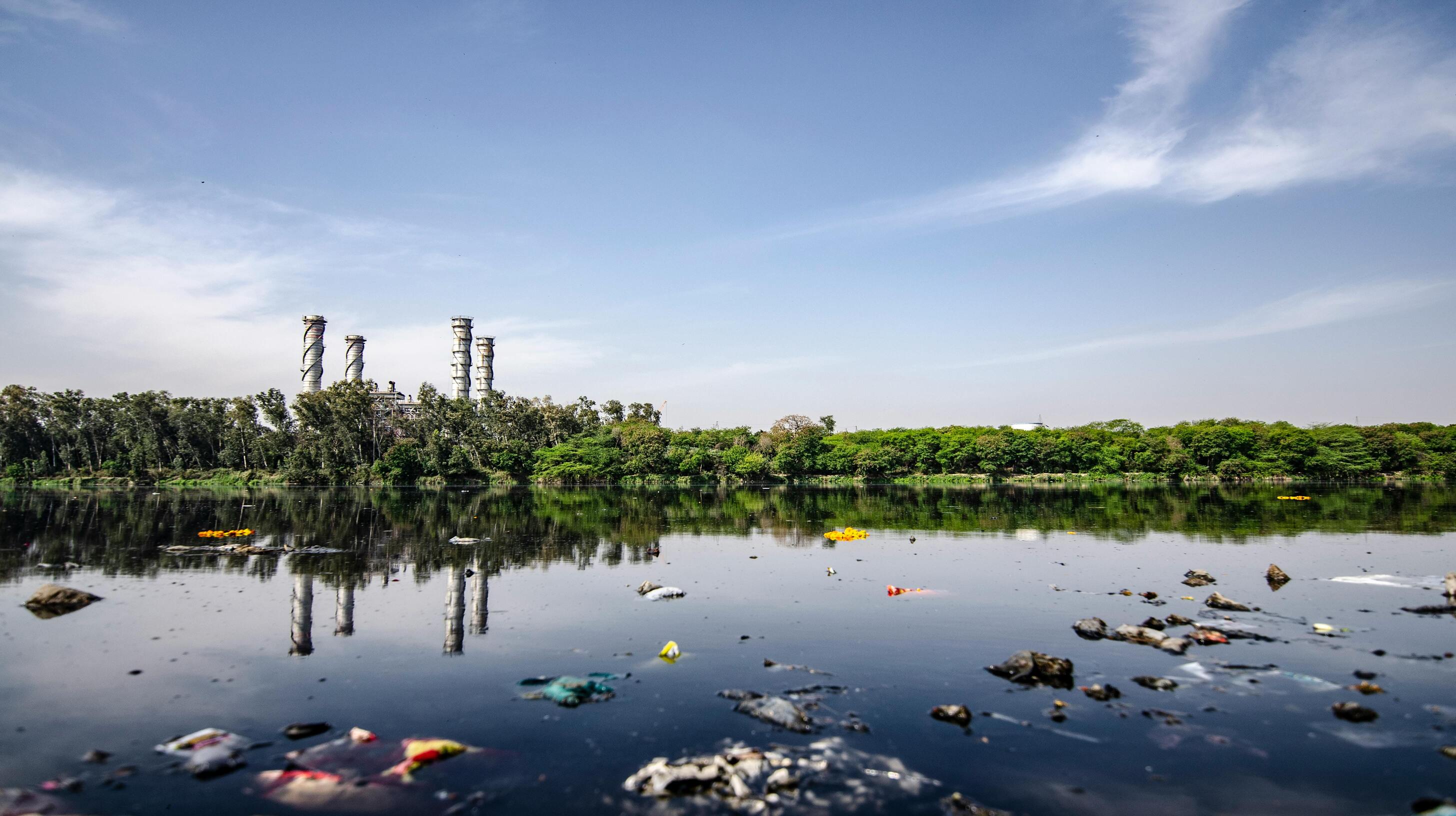 Een vervuilde rivier met drijvend afval op de voorgrond. In de achtergrond staan hoge schoorstenen van een PFAS-fabriek, omgeven door weelderige bomen. De lucht is helder met enkele wolken.